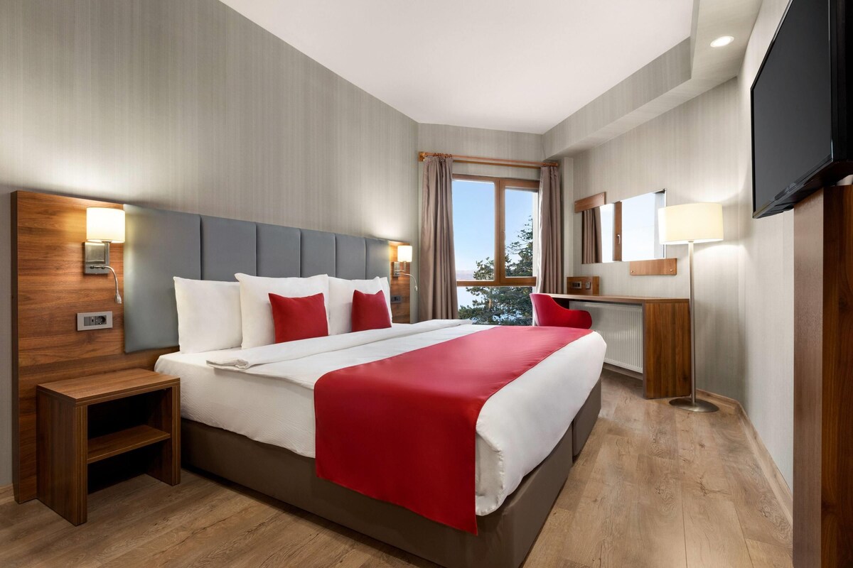 Habitación de hotel con una cama grande, cabecera de madera, mesitas de noche, televisor y una ventana con cortinas.