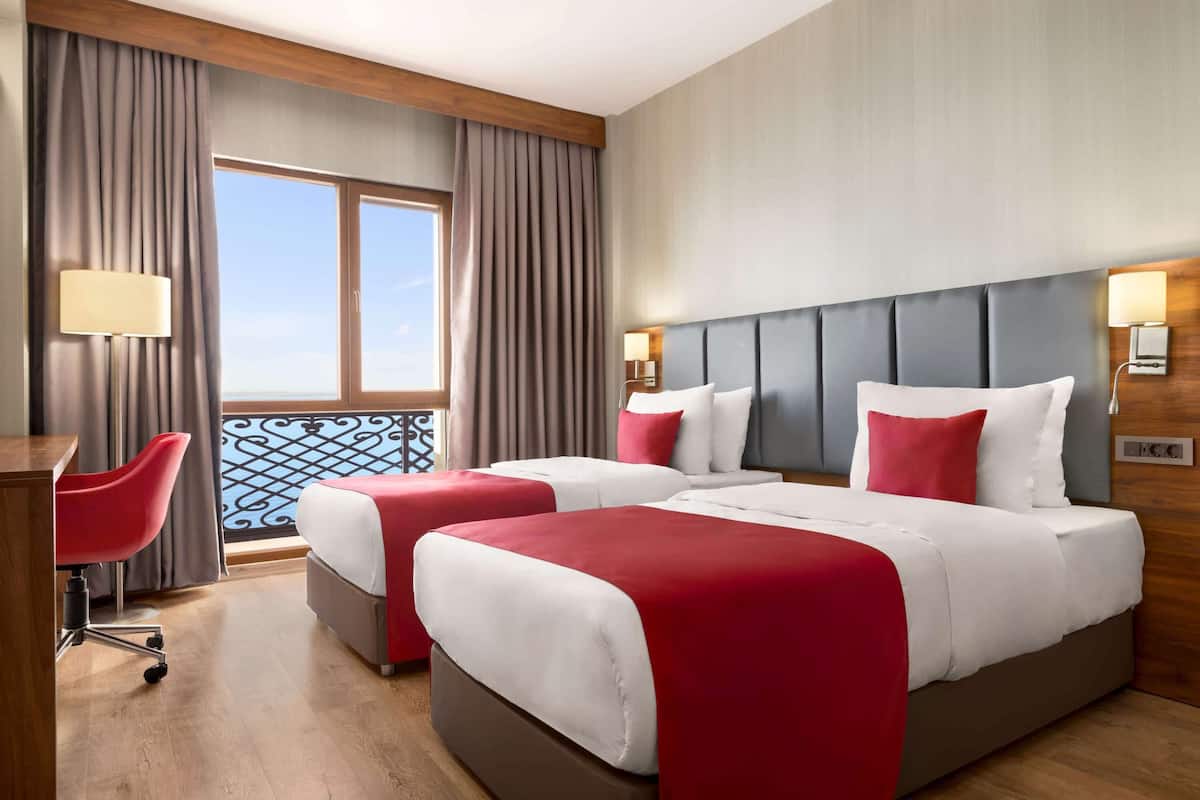 A hotel room with two beds, a desk, a chair, and a view of the sea.