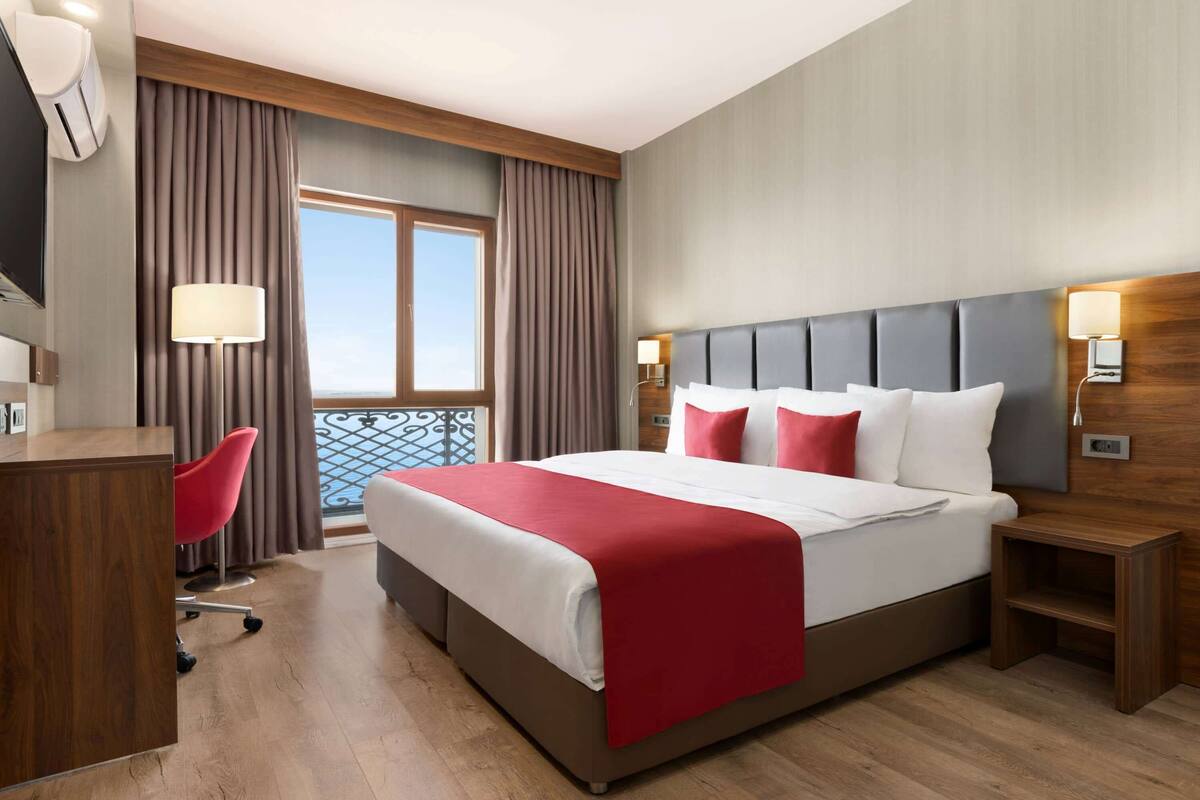 A hotel room with a large bed, a desk, a chair, a window with curtains, and a balcony view.