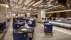 Een modern restaurantgedeelte in een hotel, met blauwe stoelen en tafels, een buffet met diverse gerechten, en een televisie die aan het plafond is bevestigd.