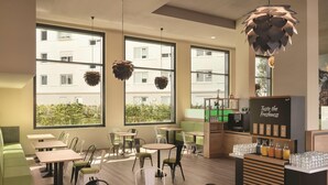 L’intérieur d’un café, avec des tables et des chaises, de grandes fenêtres et un présentoir de boissons.