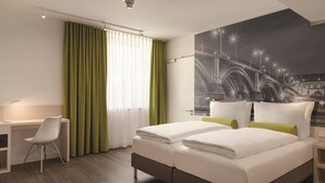 Une chambre d’hôtel avec un lit, un bureau, une chaise et une grande fenêtre avec des rideaux.
