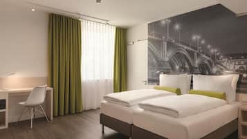 Una habitación de hotel con una cama, un escritorio, una silla y un ventanal con cortinas.