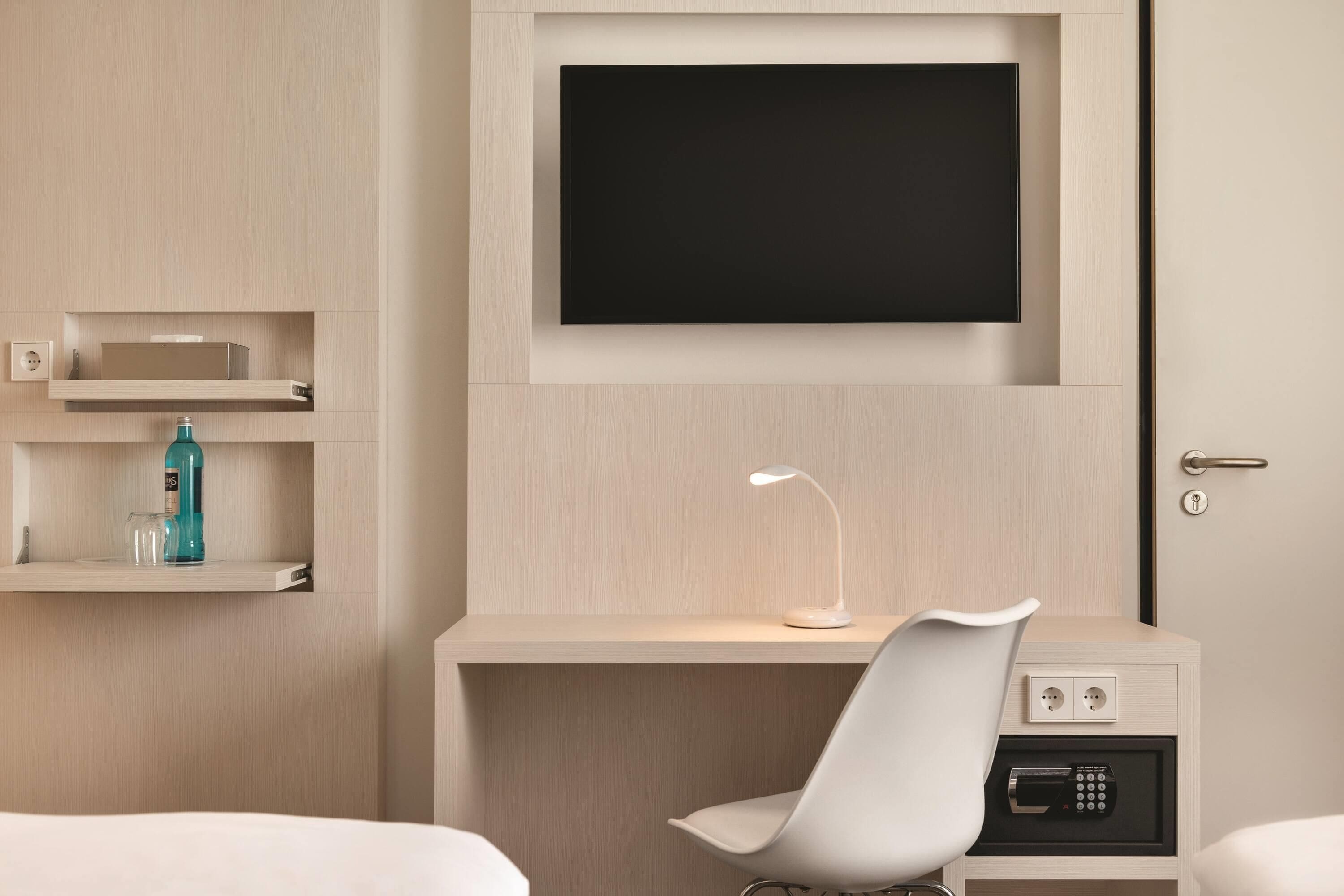 Una habitación de hotel con una cama, un escritorio con televisor, una silla, una lámpara y un panel de control.