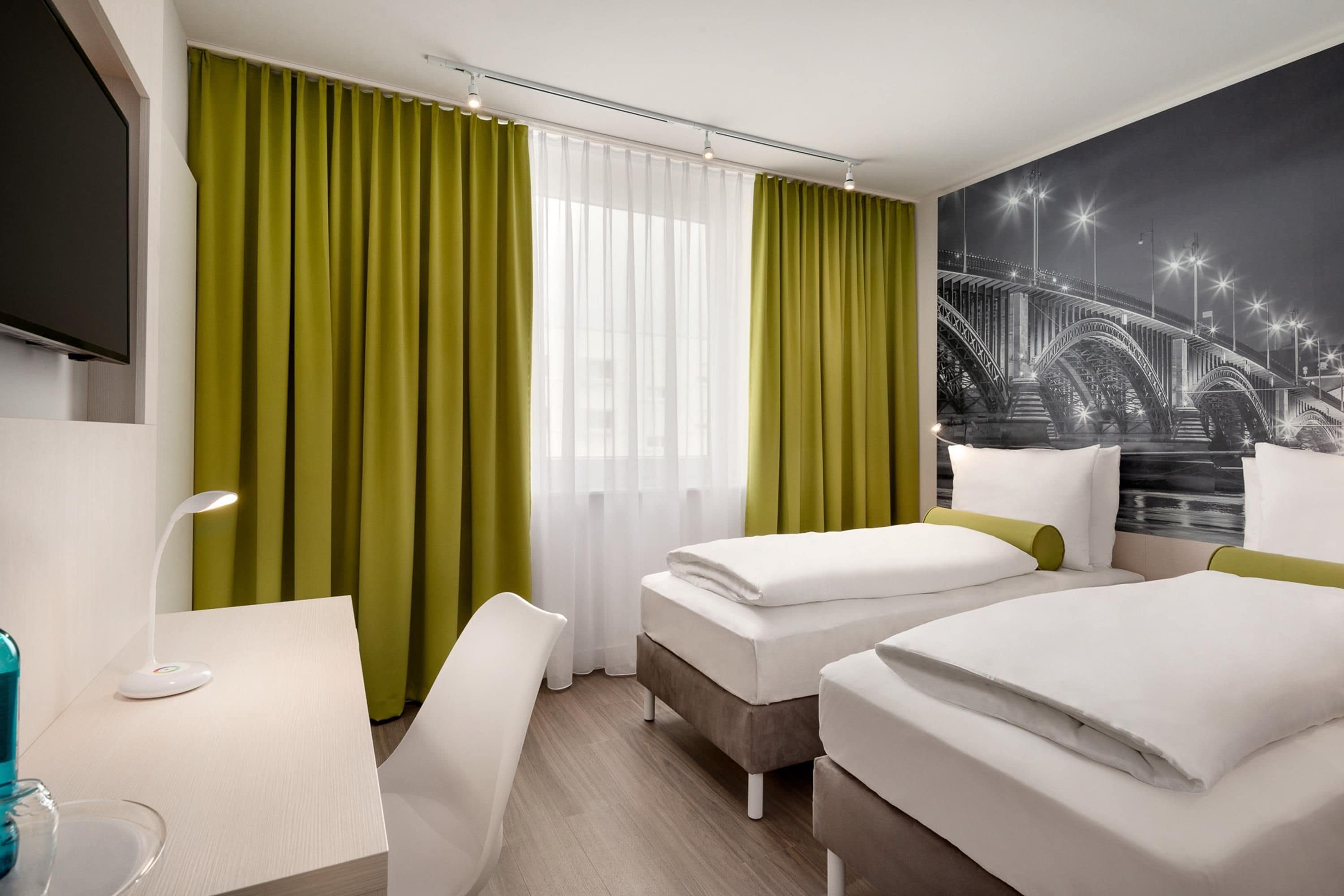 Habitación de hotel con dos camas, un escritorio, una silla, un televisor y un ventanal con cortinas.