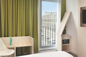 A hotel room with a bed, a desk, a chair, a bedside table with a lamp, a TV, and a view of a building through a window. - Super 8 by Wyndham Mainz Zollhafen (Mainz)