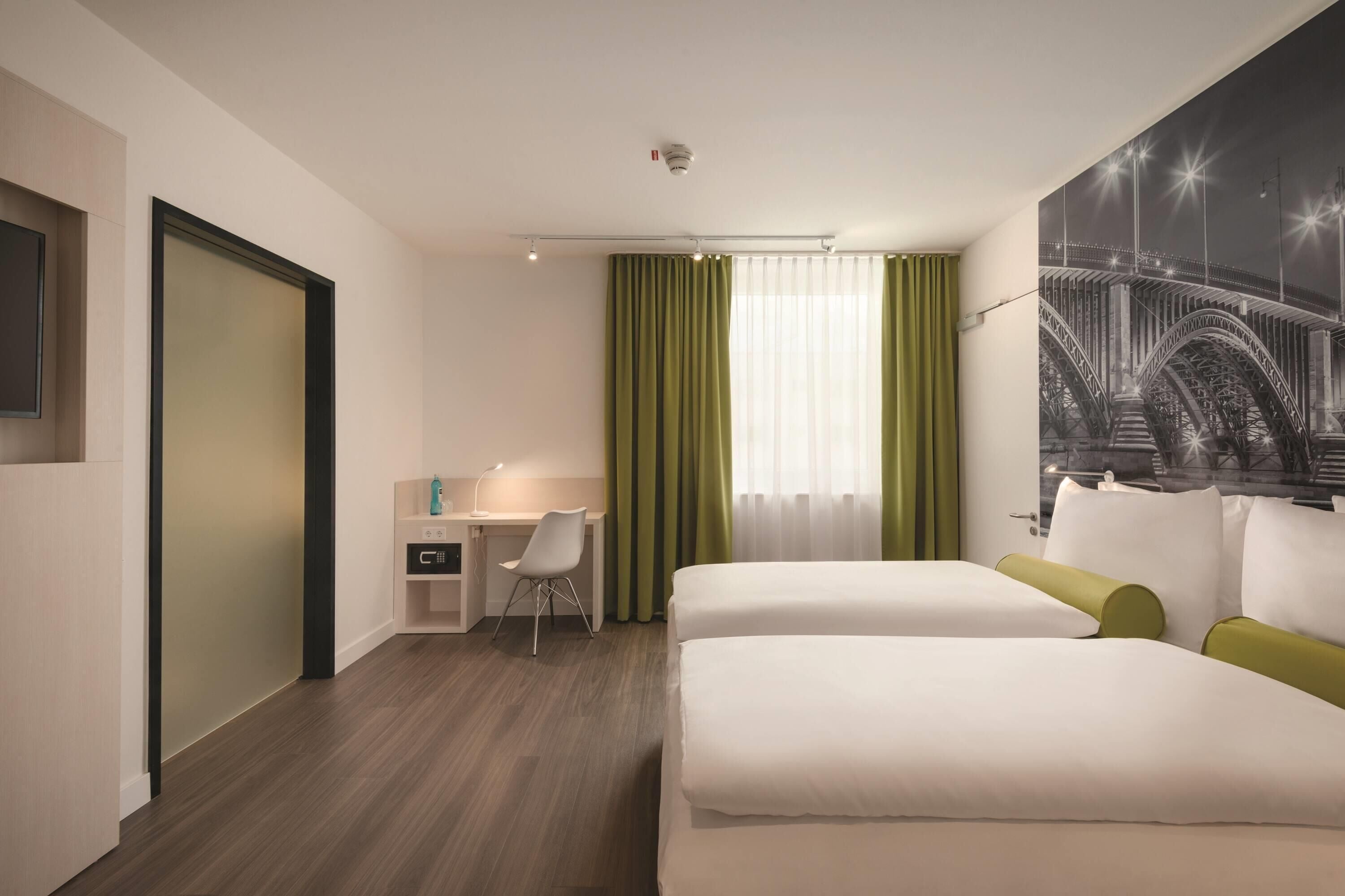 Quarto de hotel com cama, escrivaninha, cadeira e uma janela grande com cortinas.