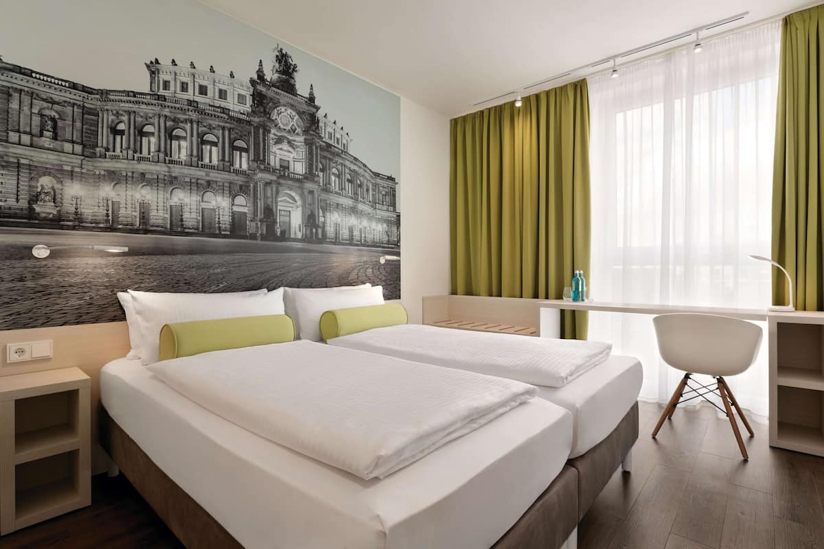 Une chambre d’hôtel avec deux lits, un bureau, une chaise et une grande fresque représentant un bâtiment historique.