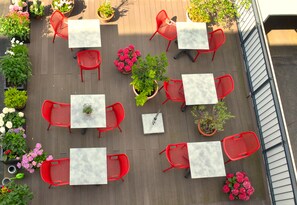 An outdoor patio with red chairs and tables, surrounded by potted plants and flowers. - Lizz Hotel Munich (Munich)