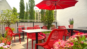 An outdoor patio with red chairs and tables, surrounded by potted plants and flowers. - Lizz Hotel Munich (Munich)