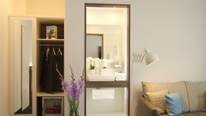 A hotel room with a walk-in closet, a bathroom with a sink and mirror, and a sofa with patterned cushions. - Lizz Hotel Munich (Munich)