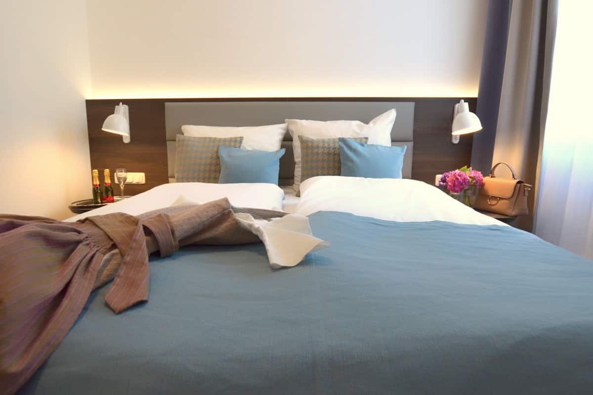 A neatly made bed with a blue bedspread, white pillows, and a brown robe on the side. There are two bedside lamps and a nightstand with a bottle and glass.