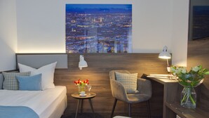 A hotel room with a bed, a chair, a small table with a vase of flowers, and a cityscape view on the wall. - Lizz Hotel Munich (Munich)