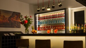 A bar area with a well-lit shelf of liquor bottles, two bar stools, a vase with flowers, and a cityscape painting on the wall. - Lizz Hotel Munich (Munich)
