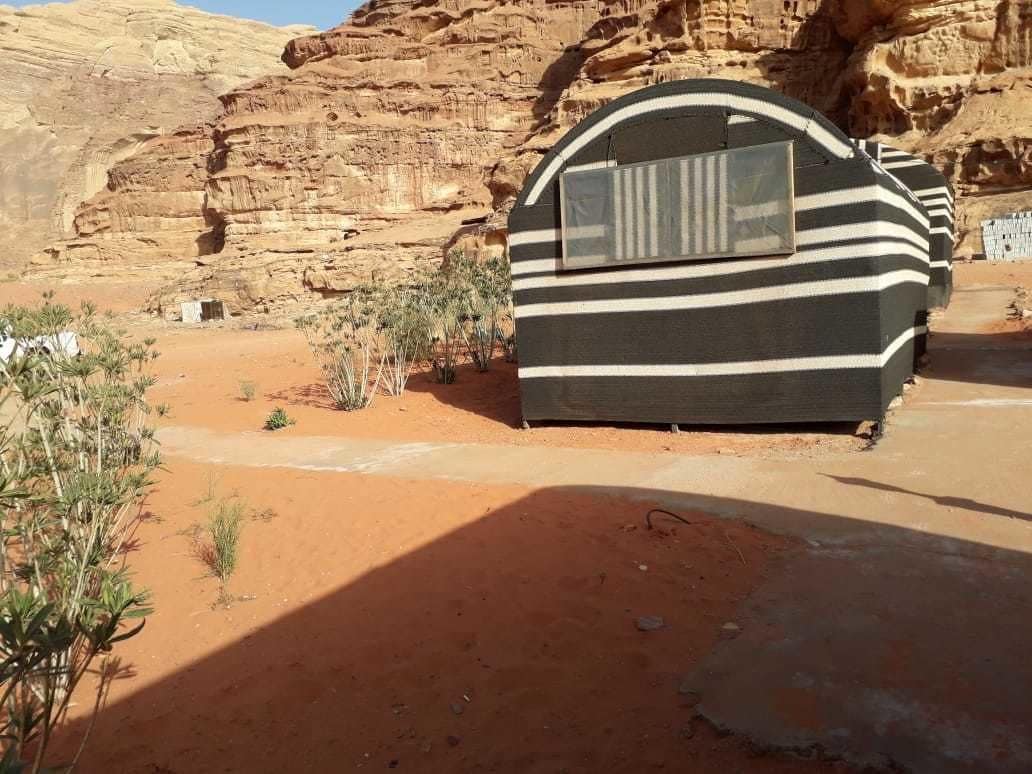A small, single-room structure with a curved roof and a window, situated in a desert environment with red sand and sparse vegetation.