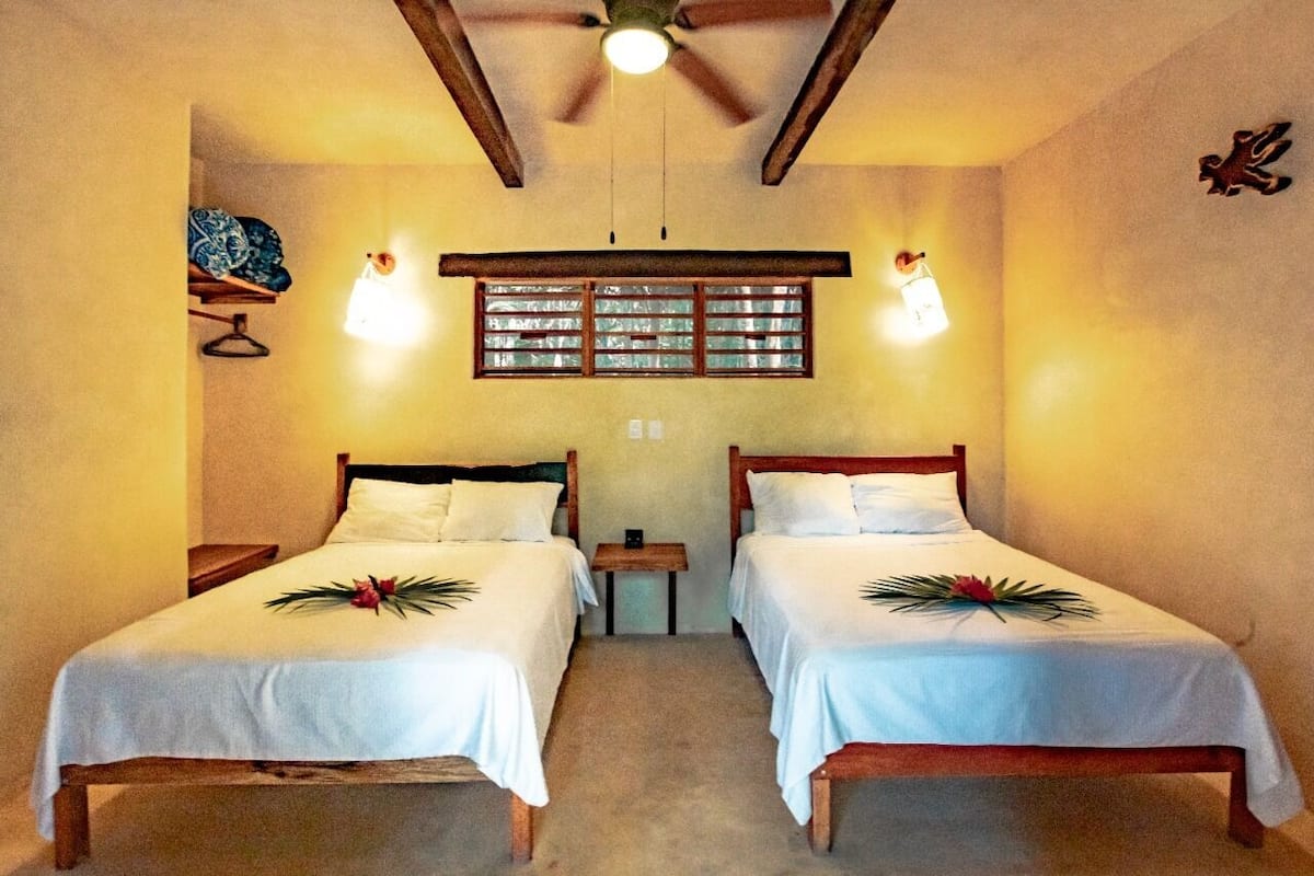 A hotel room with two single beds, a ceiling fan, a window with blinds, and a small table between the beds.