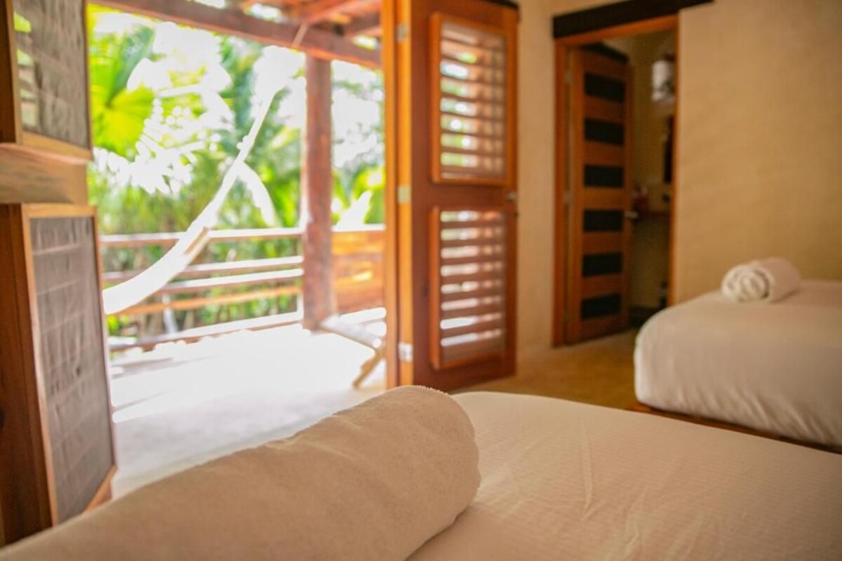 A room with two beds, a wooden door, and a view of greenery outside.