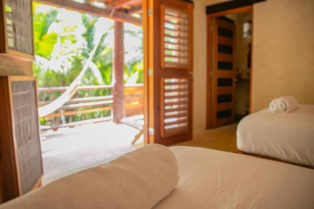 A room with two beds, a wooden door, and a view of greenery outside.