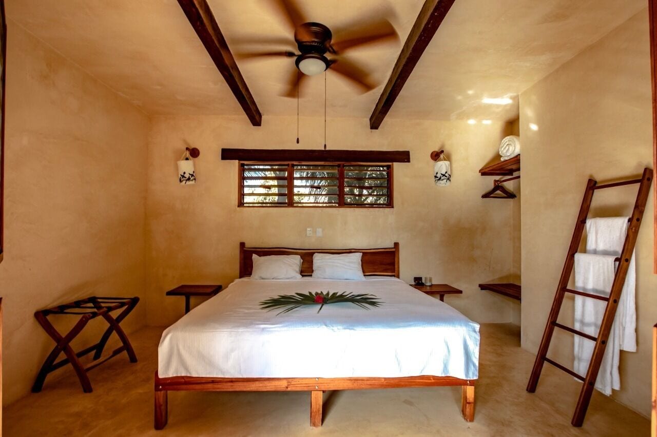 Un dormitorio con cama de madera, un ventilador de techo, una ventana con persianas, una escalera de madera y una mesita pequeña.
