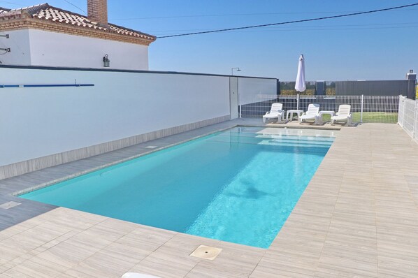 A rectangular swimming pool with a tiled deck, white lounge chairs, and a white fence.