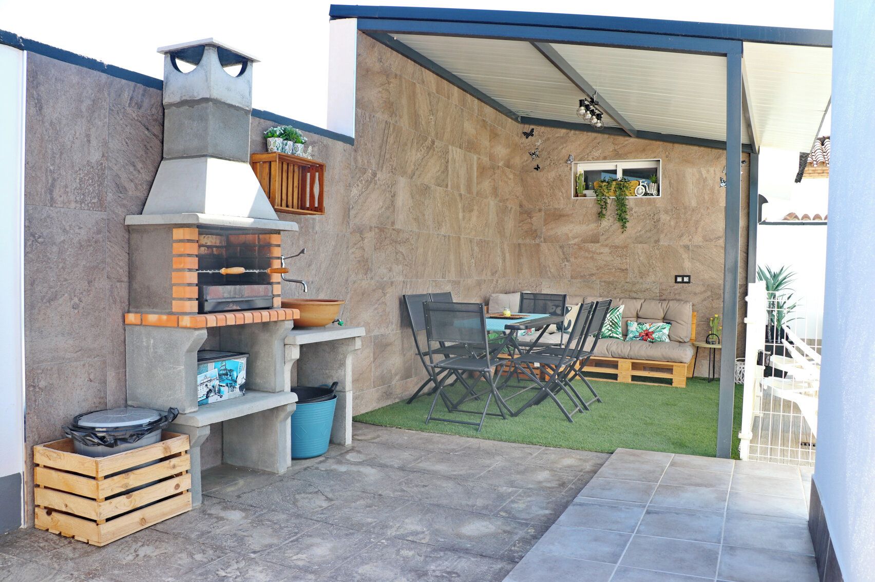 Een terras buiten met een stenen muur, een ingebouwde barbecue, een eettafel met stoelen en een klein zitgedeelte met kussens.