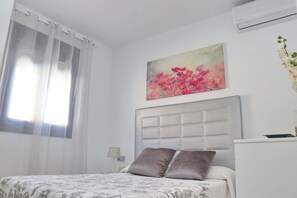 A bedroom with a bed, a headboard, a painting, a window with curtains, and an air conditioning unit.