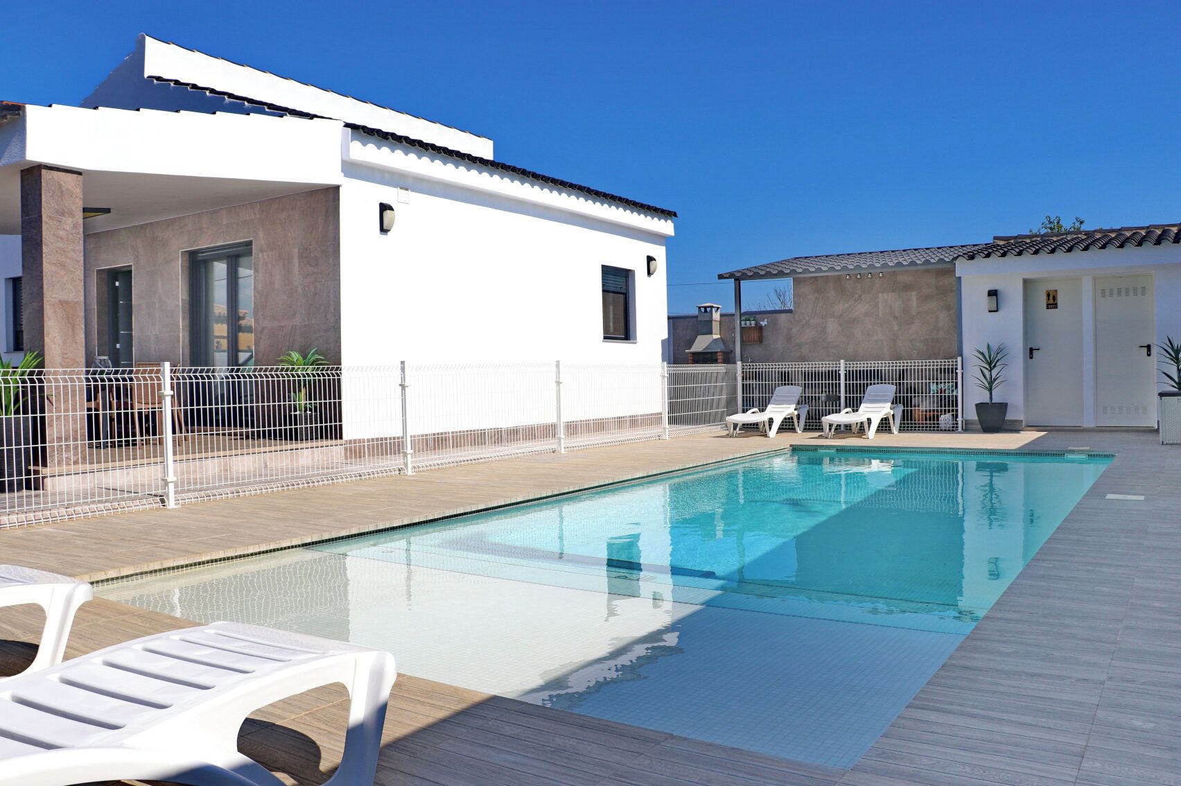 A modern outdoor swimming pool with a wooden deck, white lounge chairs, and a white building with large windows.