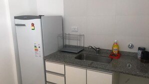 Cocina compacta con refrigerador, pileta y estante metálico.