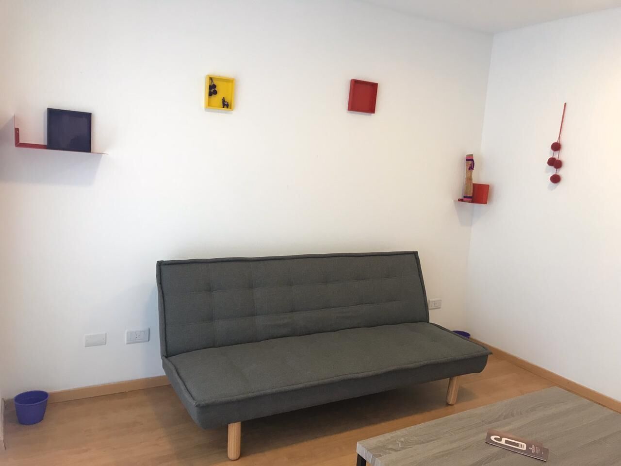 A minimalist room with a grey sofa, a small blue trash bin, and a wooden coffee table.