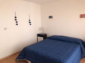A bedroom with a blue bedspread, a small bedside table with a lamp, and wall decorations including a string of spherical objects and a small framed picture.