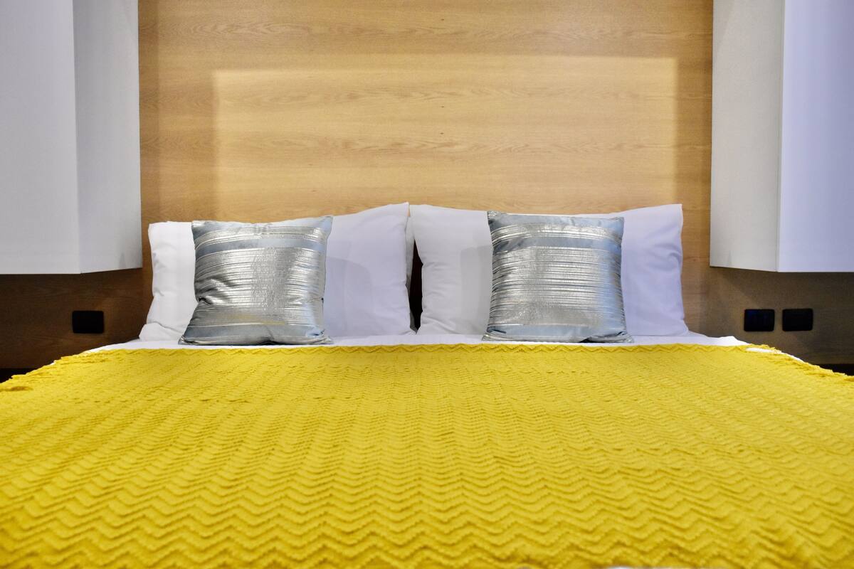 A bed with a yellow textured bedspread, two white pillows, and two silver pillows against a wooden headboard.