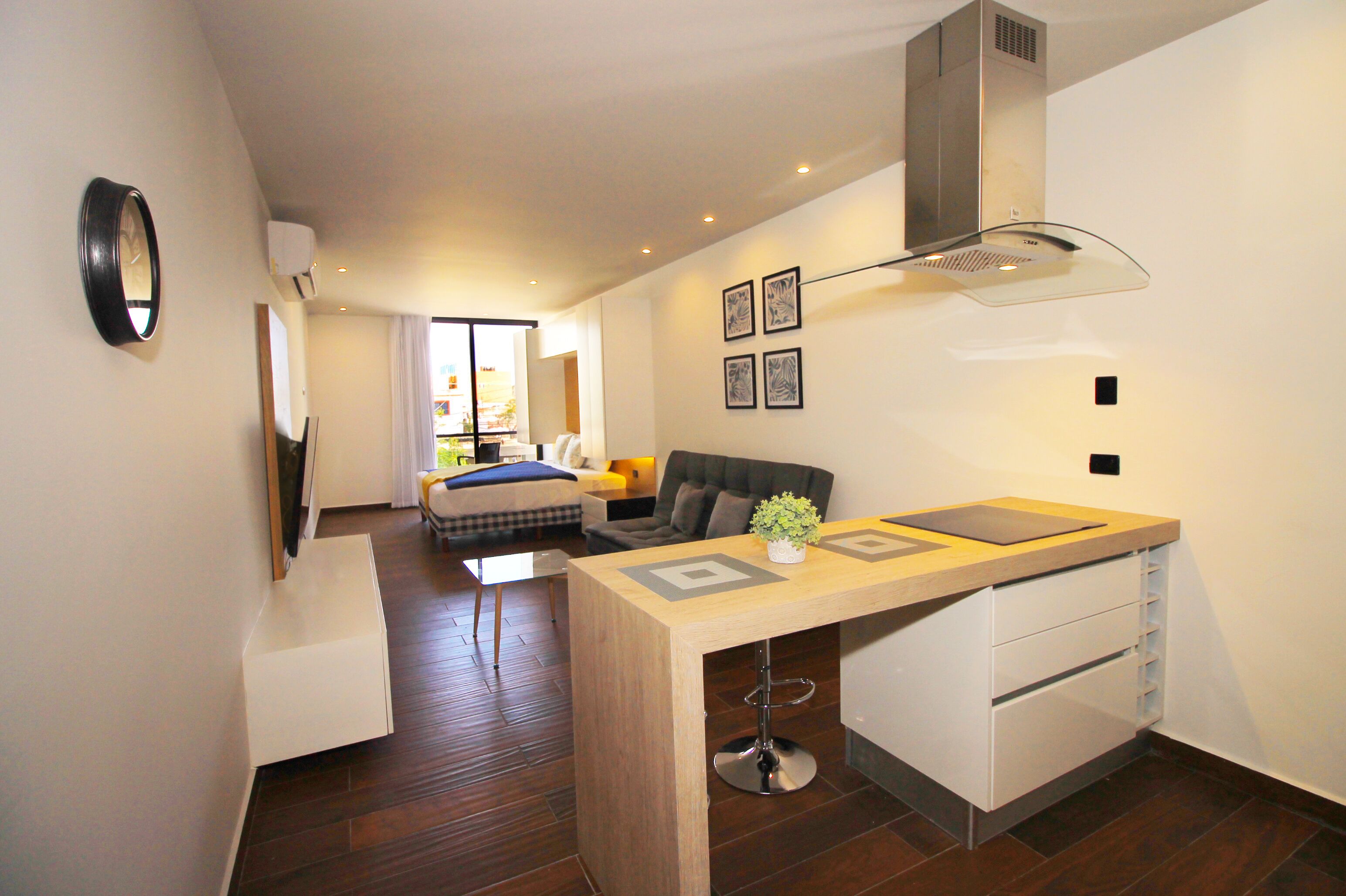 Eine moderne Küche mit Kochinsel, ein Essbereich mit Sofa und kleinem Tisch sowie ein Schlafzimmer mit Bett und Fenster.