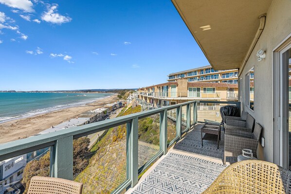 A balcony with a view of the beach, outdoor furniture, and a building with multiple windows. - Two Bedroom Condo With Great Ocean Views! (Aptos)