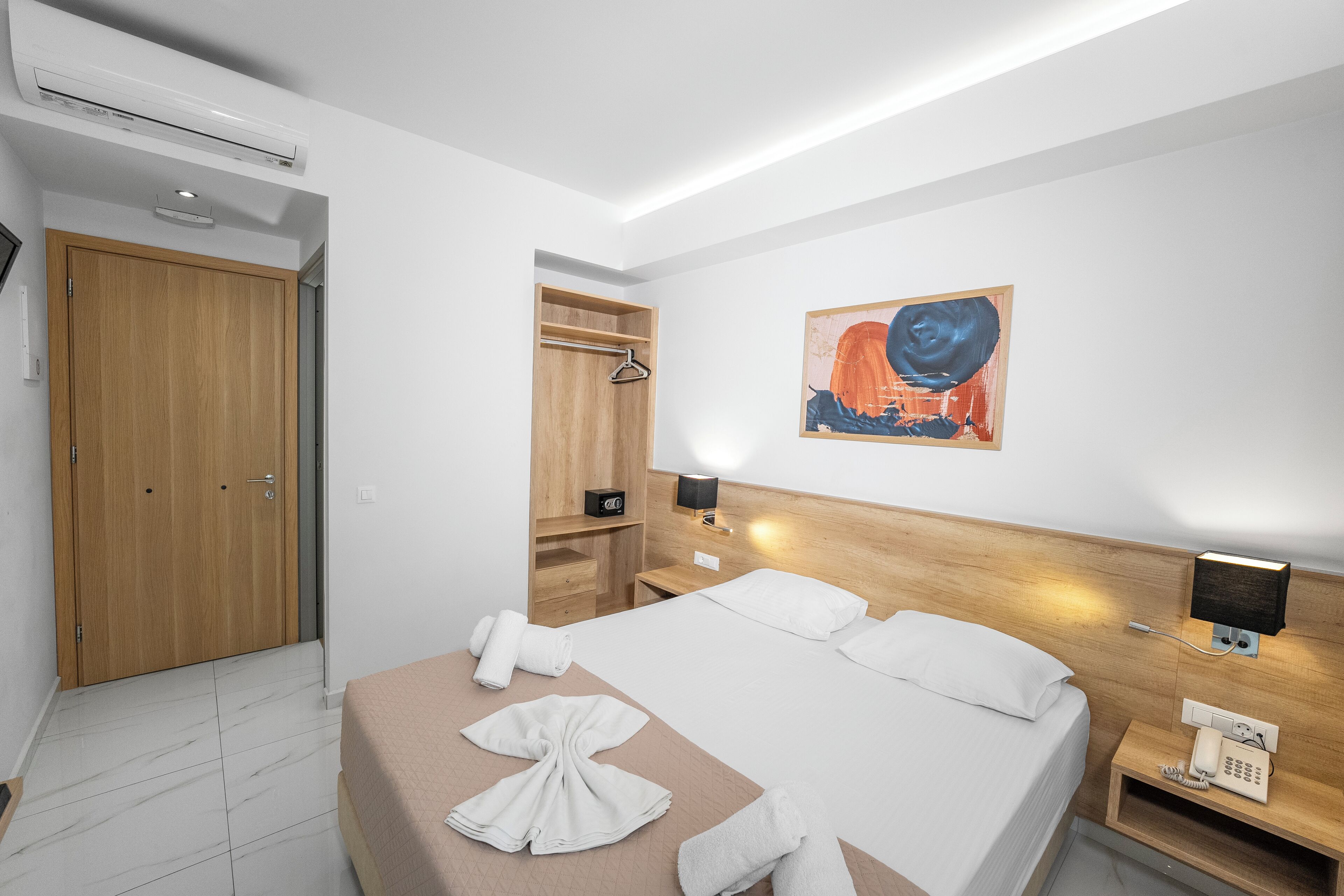 A hotel room with a bed, a wooden wardrobe, a bedside table with a lamp, a phone, and a painting on the wall.