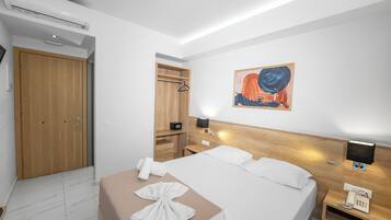 A hotel room with a bed, a wooden wardrobe, a bedside table with a lamp, a phone, and a painting on the wall.