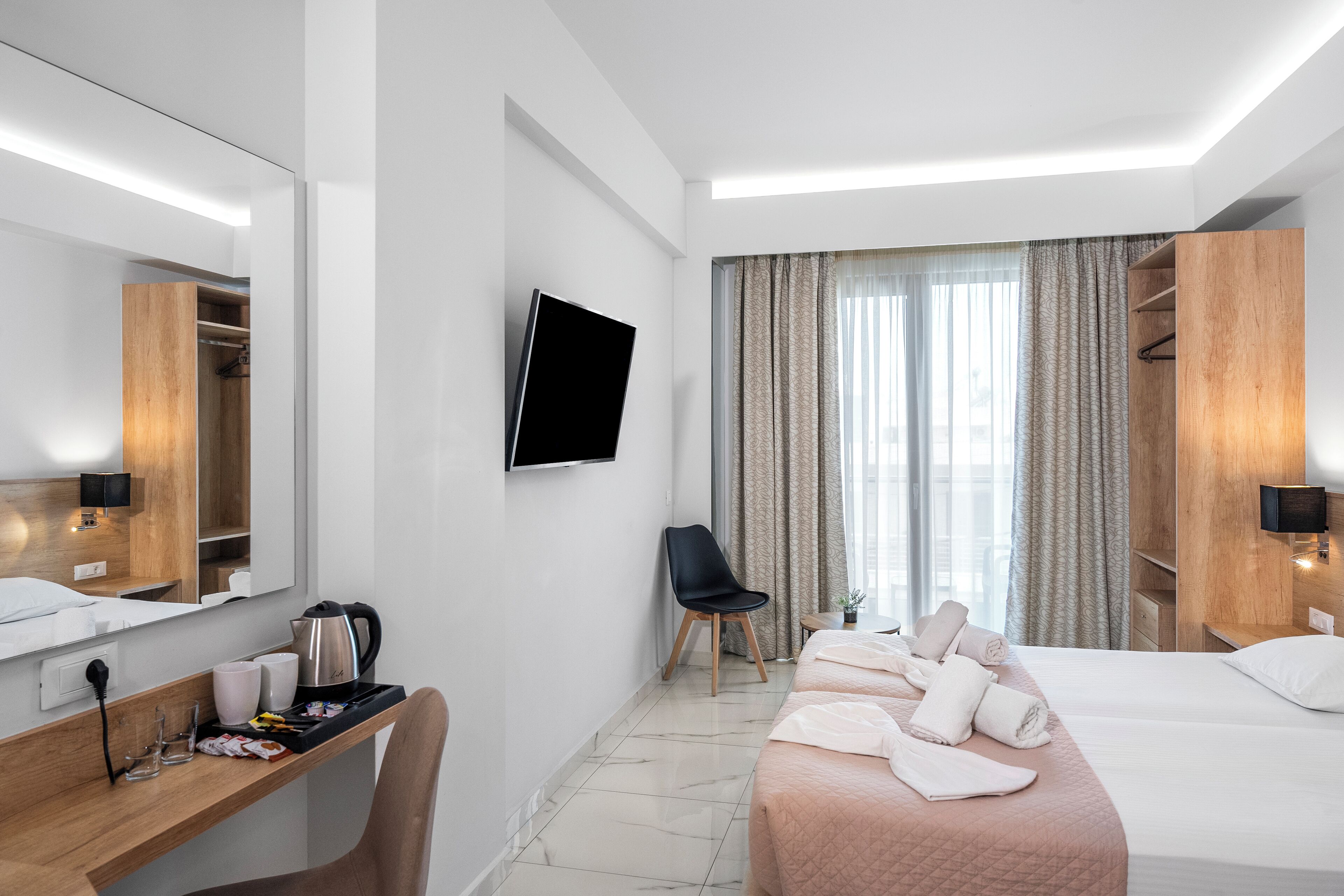 Une chambre d’hôtel moderne équipée d’un lit, d’un bureau avec une bouilloire, d’une chaise, d’une télévision et d’une grande fenêtre avec des rideaux.
