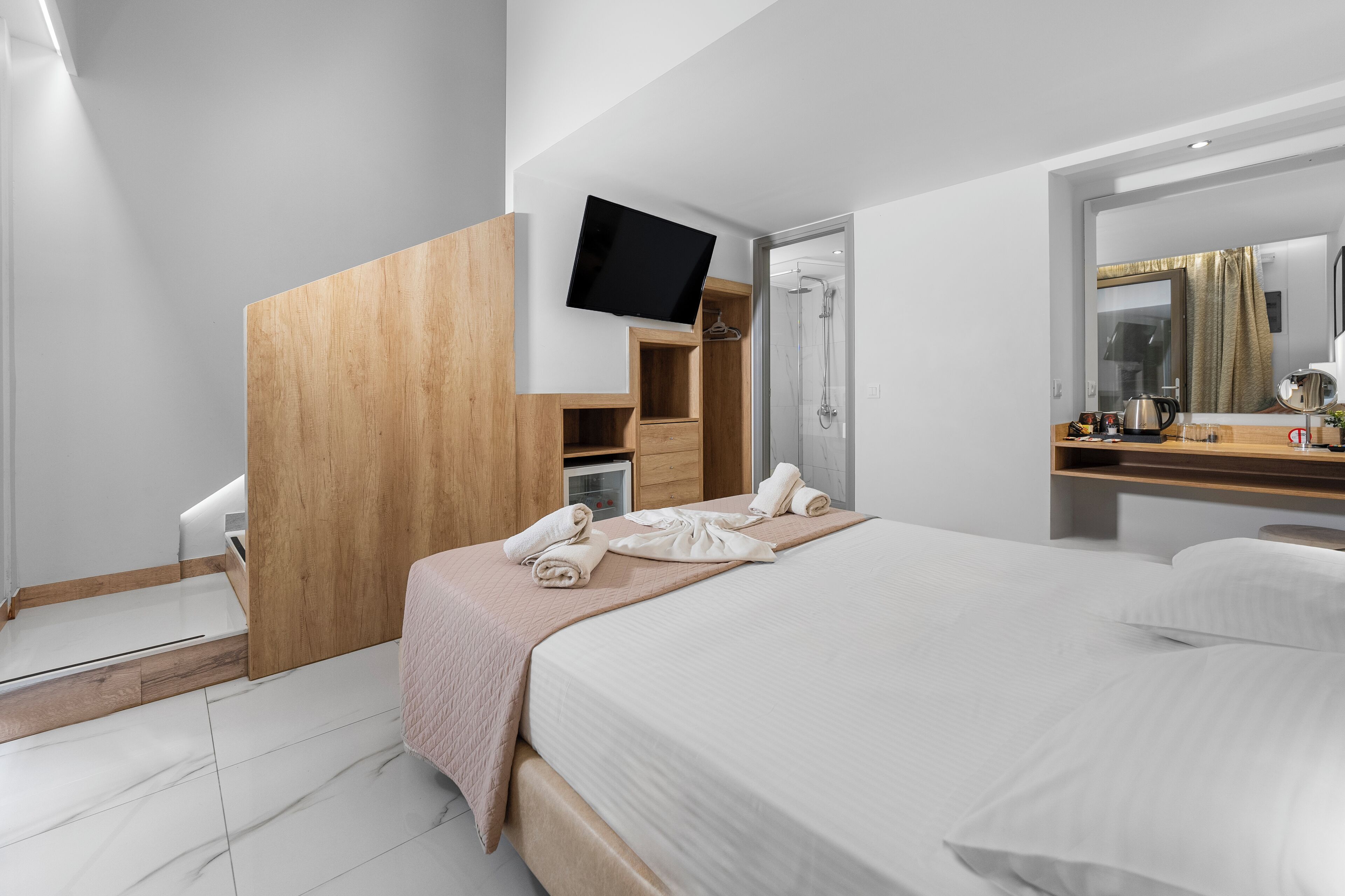 A modern hotel room with a bed, a TV mounted on a wooden panel, a shower area, and a mirror above the sink.
