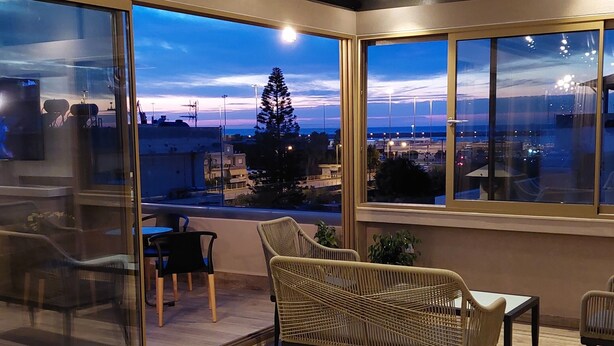 Un área de estar al aire libre con muebles de mimbre, amplias ventanas que ofrecen vistas a la ciudad e iluminación empotrada en el techo.