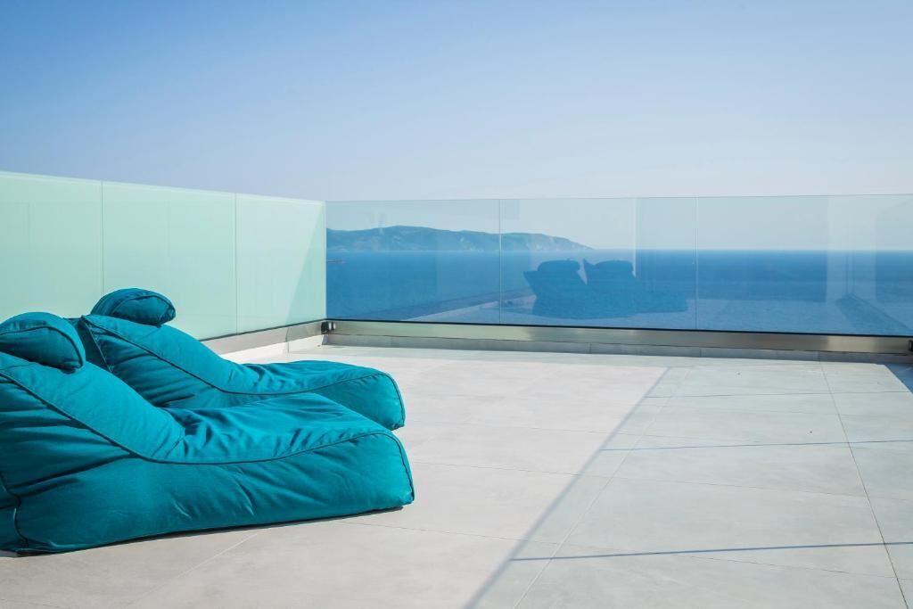 A modern balcony with a glass railing, a teal bean bag, and a clear view of the sea.