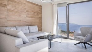 A modern living room with a sofa, armchair, and a table, large sliding glass doors leading to a balcony with a view. - Oikos Boutique Residences Private Estate (Kefalonia)