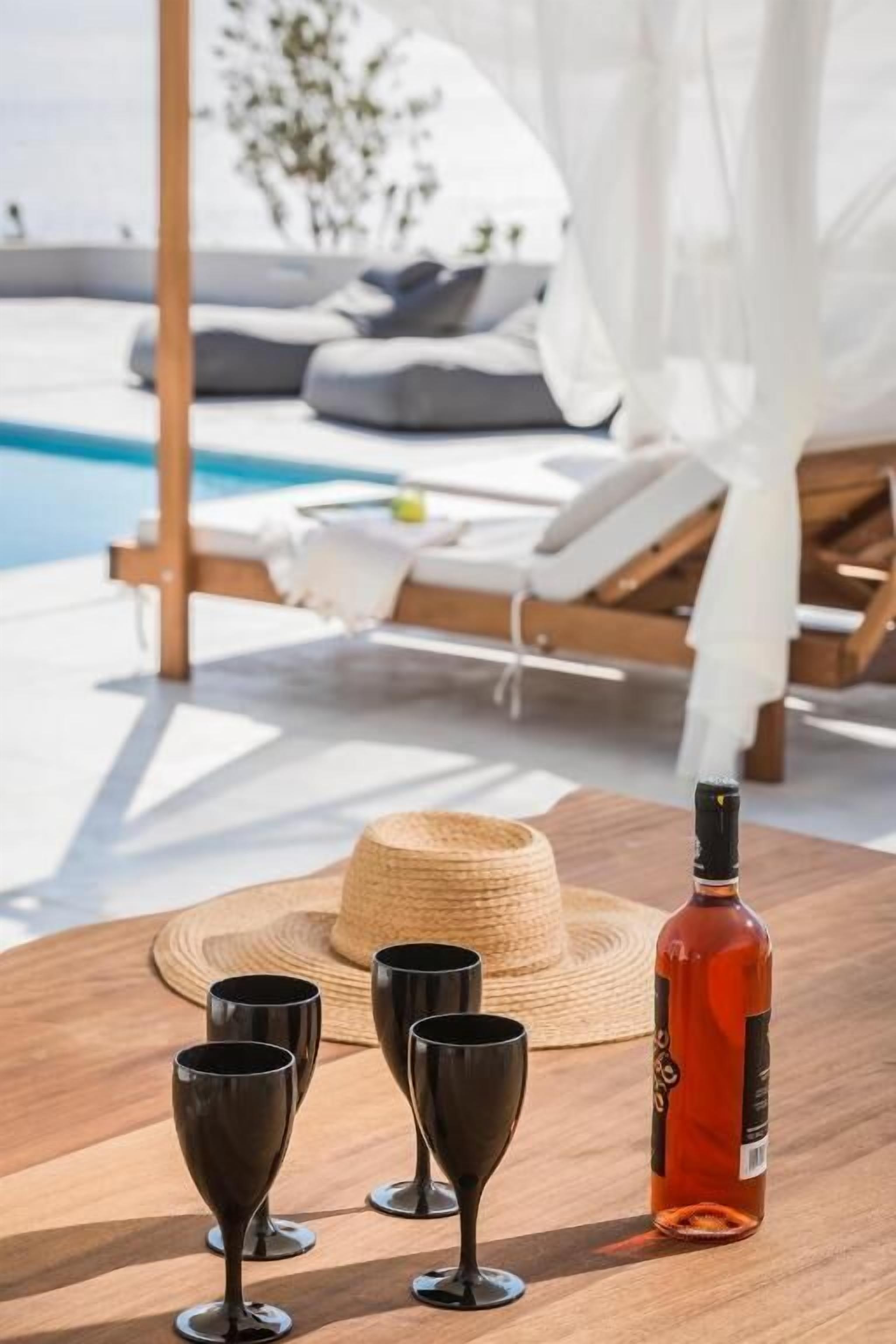 Een houten tafel met een fles en glazen, een strohoed, en een terras bij het zwembad met witte luifels en kussens.
