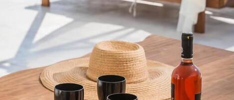 Een houten tafel met een fles en glazen, een strohoed, en een terras bij het zwembad met witte luifels en kussens.