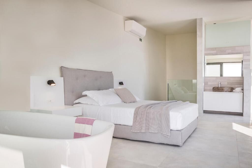 A modern bedroom with a bed, bedside tables, a glass shower enclosure, and a white bathtub.