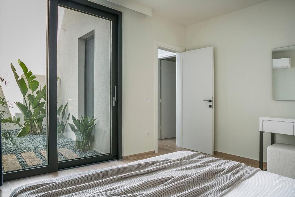 A bedroom with a bed, a sliding glass door to a garden, a white bedside table, and a door leading to another room.