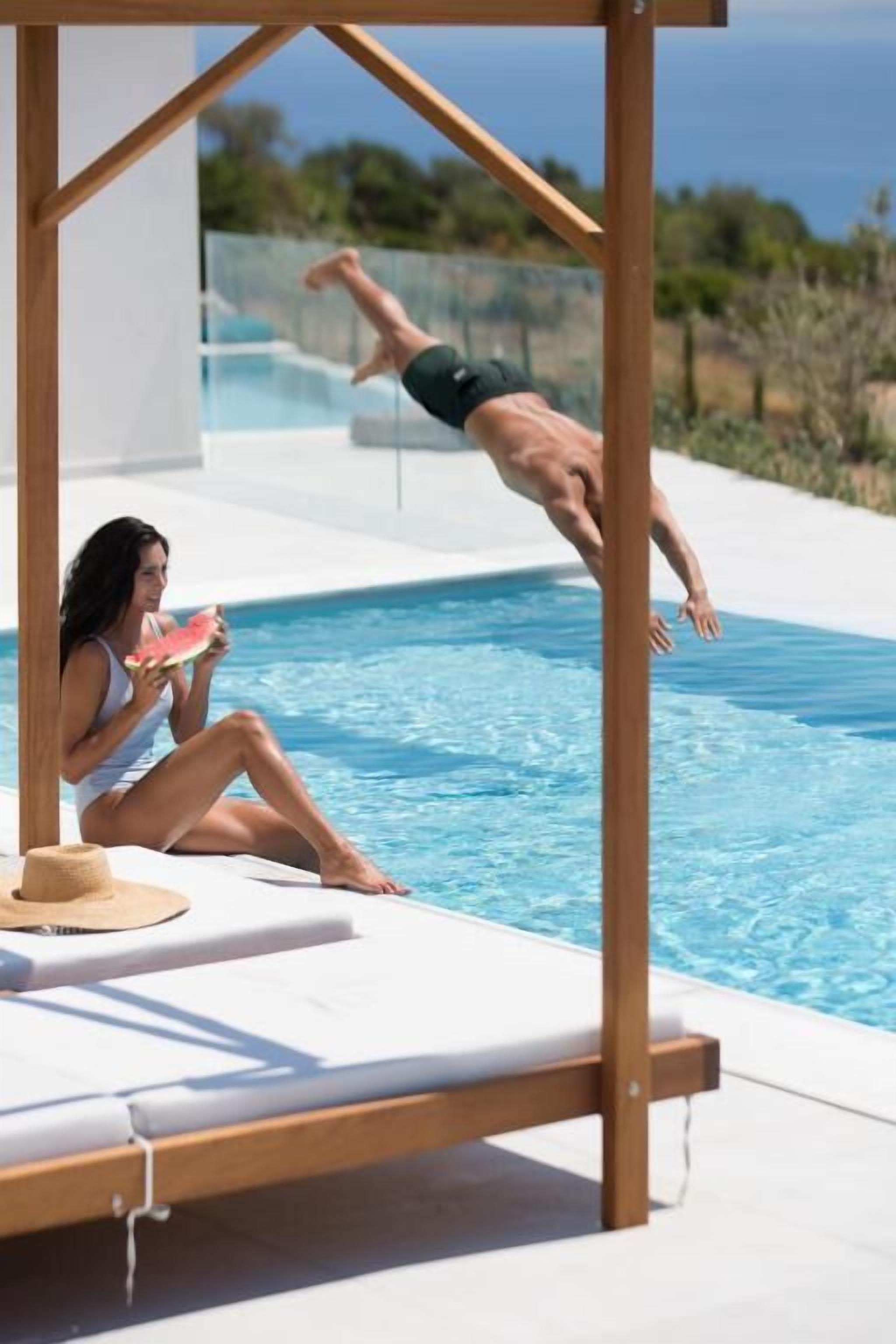 A poolside area with a diving board, a lounge chair, and a person sitting by the pool.
