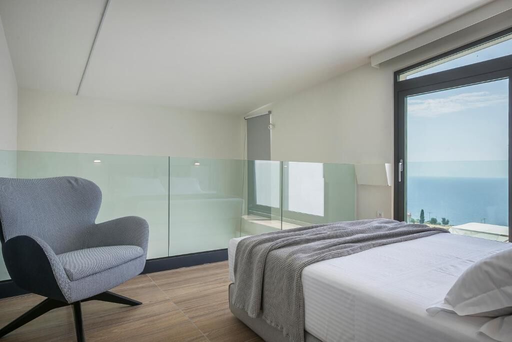Un dormitorio moderno con cama, una silla, un separador de vidrio y un balcón con vistas al mar.
