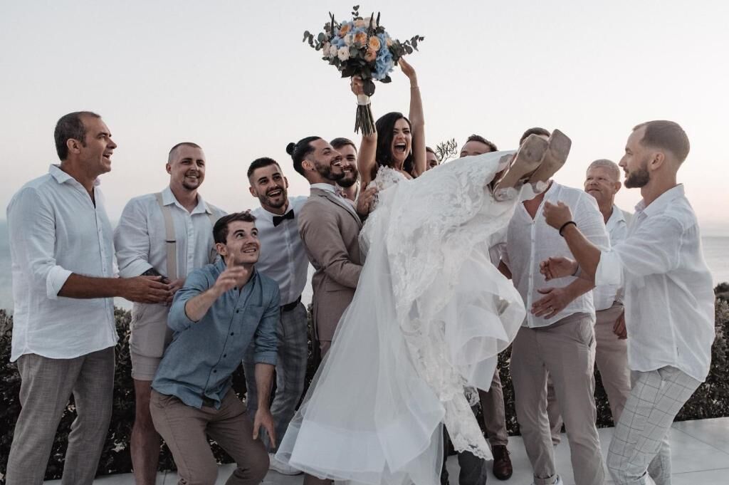A group of people outdoors, some in formal attire, with a person in a white dress holding a bouquet.