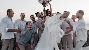 A group of people outdoors, some in formal attire, with a person in a white dress holding a bouquet.