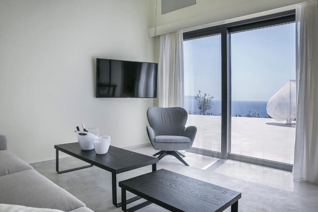 A modern living room with a flat-screen TV, a grey armchair, a coffee table, and a balcony with a view of the sea.