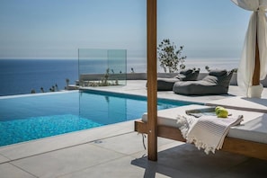 A modern infinity pool with a clear view of the ocean, a wooden pergola, and a cozy seating area with cushions and a blanket.